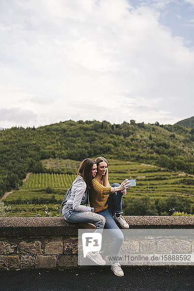 Zwei junge Frauen sitzen auf einer Mauer in ländlicher Landschaft und machen einen Selfie  Greve in Chianti  Toskana  Italien