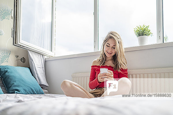 Smiling young woman sitting on bed at home using cell phone