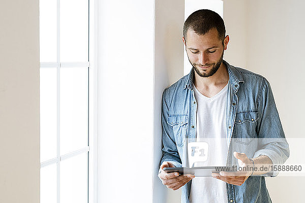 Young man at home smiling and looking at his tablet standing at window