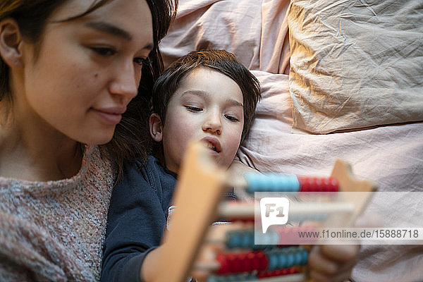 Mother and little son lying together on bed using abacus
