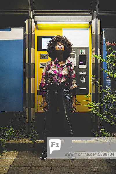 Junge Frau mit Afrofrisur steht nachts am Fahrkartenautomaten und schaut auf