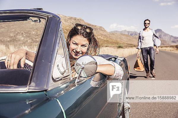 Woman in convertible car on a road trip waiting for man with a travelling bag