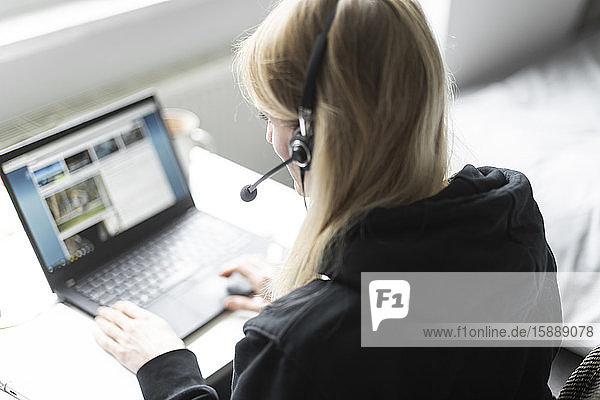 Blonde Frau mit Kopfhörer sitzt zu Hause am Schreibtisch und arbeitet am Laptop