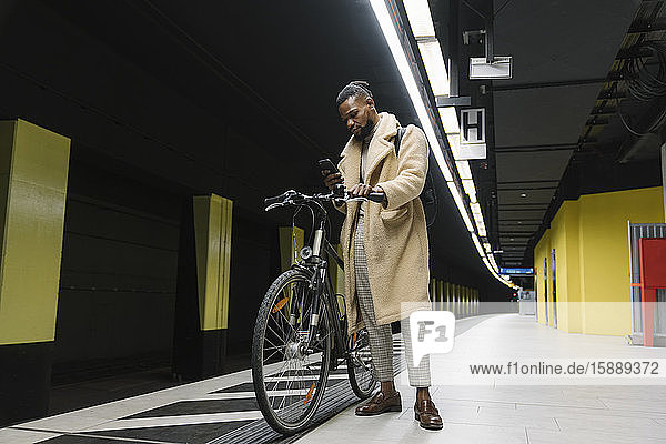 Stilvoller Mann mit Fahrrad und Smartphone wartet auf die U-Bahn