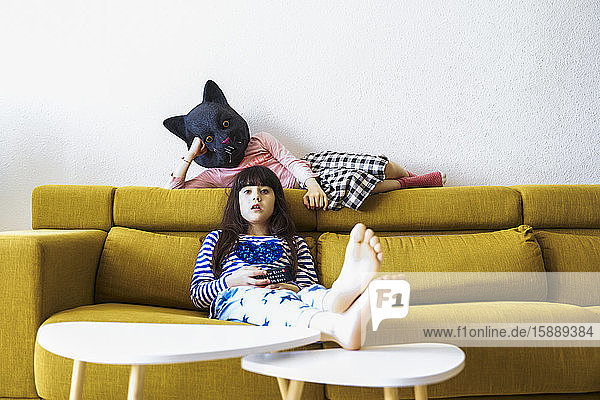 Zwei gelangweilte Mädchen sitzen auf der Couch  sehen fern  eine trägt eine Katzenmaske