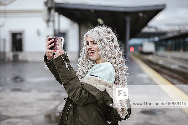 Porträt einer lachenden jungen Frau  die sich mit einem Smartphone auf dem Bahnsteig selbstständig macht