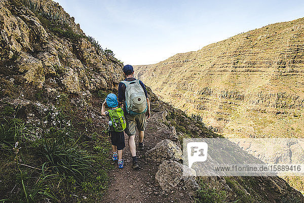 Back view of father and little son with backpacks walking on a hiking trail in the mountains  La Gomera  Canary Islands  Spain