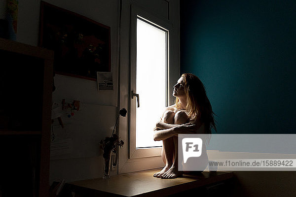 Seriöse junge Frau sitzt zu Hause auf der Fensterbank und schaut hinaus