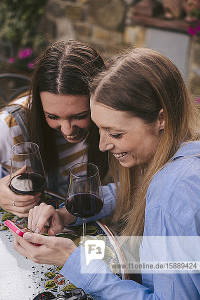 Zwei glückliche Frauen  die ein Glas Rotwein trinken und im Freien auf ihr Handy schauen