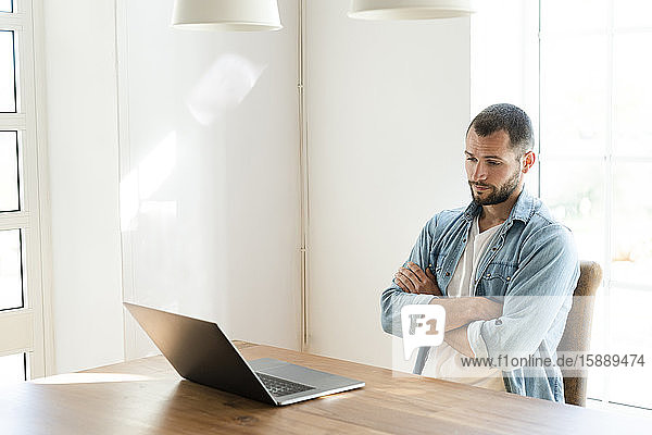 Schwerer junger Mann mit verschränkten Armen zu Hause  der im Heimbüro auf seinen Laptop schaut