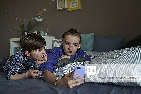 Portrait eines Bruders und seiner älteren Schwester  die zusammen auf dem Bett liegen und ein Smartphone für Video-Chat benutzen