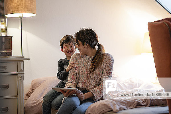 Mutter und kleiner Sohn sitzen gemeinsam mit einem digitalen Tablet am Bett und haben Spaß