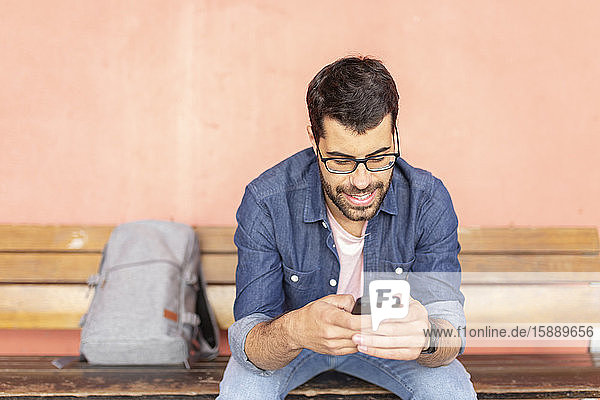 Porträt eines lächelnden Mannes  der auf einer Holzbank sitzt und ein Smartphone benutzt