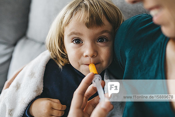 Porträt eines kranken kleinen Mädchens  das auf einer Couch sitzt  während die Mutter ihr Medikamente gibt