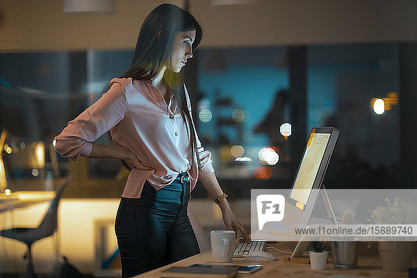 Serious young woman standing at desk in office looking at computer
