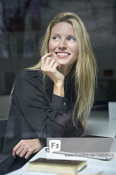 Porträt einer glücklichen blonden Frau mit Laptop hinter einer Fensterscheibe im Büro