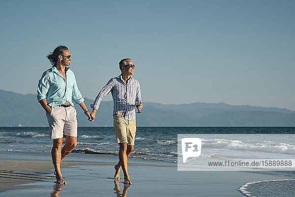 Lächelnde reife schwule Männer in voller Länge  die sich an den Händen halten  während sie am Strand vor klarem blauen Himmel an der Küste spazieren gehen  Riviera Nayarit  Mexiko