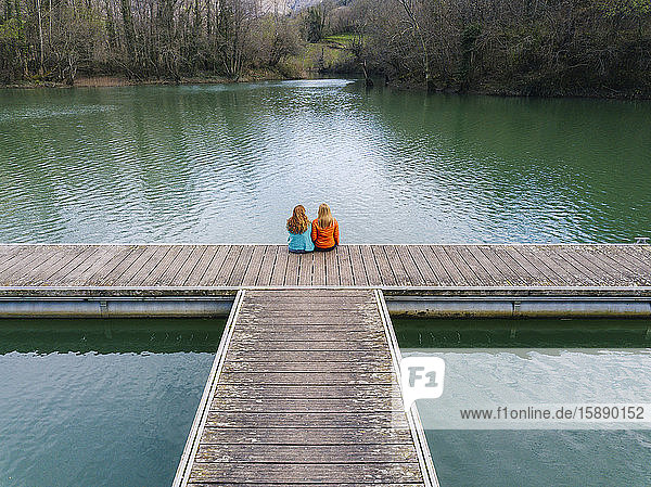 Rückenansicht von zwei Freunden  die nebeneinander auf dem Steg sitzen  Valdemurio-Stausee  Asturien  Spanien