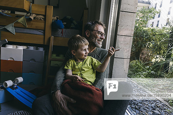 Vater und kleiner Sohn sitzen zu Hause auf dem Boden und schauen aus dem Fenster