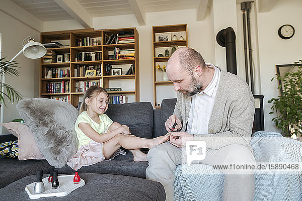 Father painting his daughter's toenails with red nail polish on the couch in living room