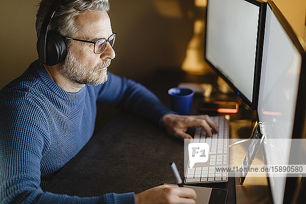 Älterer Mann mit Kopfhörern sitzt zu Hause am Schreibtisch und arbeitet mit Grafiktablett und Computer
