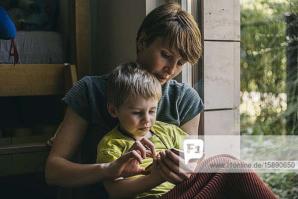 Mutter und kleiner Sohn kuscheln zu Hause und schauen auf das Smartphone
