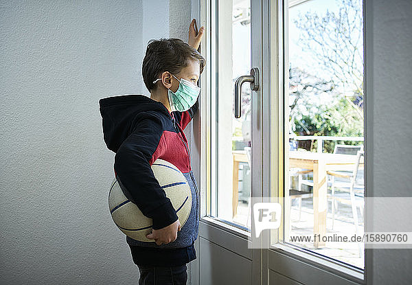 Trauriger Junge mit Basketball und Maske  der aus dem Fenster schaut