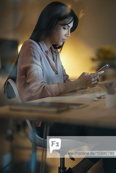 Junge Frau sitzt im Büro am Schreibtisch und schaut auf ein Smartphone