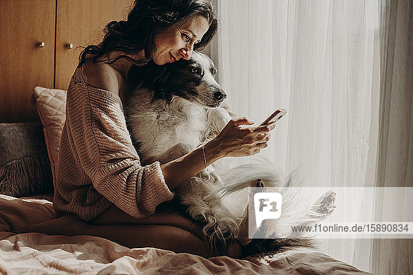 Lächelnde Frau sitzt mit ihrem Hund auf dem Bett und nimmt Selfie mit Smartphone
