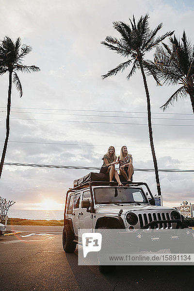 Freundinnen in voller Länge im Jeep vor Palmen und bewölktem Himmel sitzend  USA  Hawaii  Maui