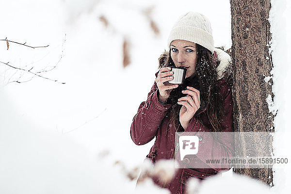Porträt einer jungen Frau  die sich im Winter an einen Baumstamm lehnt und ein heisses Getränk trinkt