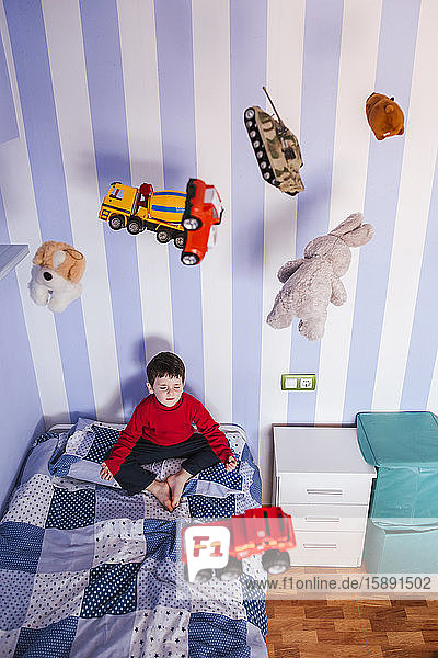 Kleiner Junge meditiert auf seinem Bett mit Spielzeug  das über seinen Kopf fliegt