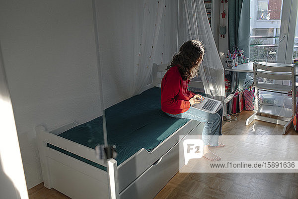 Mädchen mit Laptop sitzt auf dem Bett im Kinderzimmer und schaut aus dem Fenster