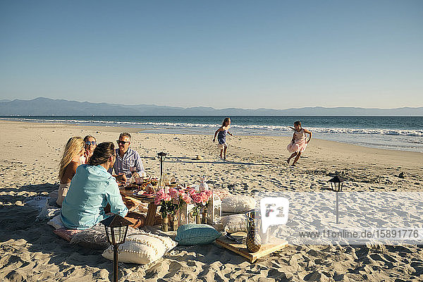 Ältere männliche und weibliche Freunde genießen das Picknick  während die Mädchen am Strand bei strahlendem Sonnenschein gegen den klaren Himmel spielen. Riviera Nayarit  Mexiko