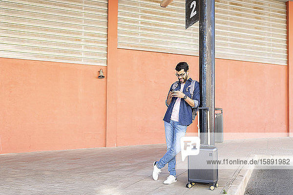 Mann mit Rucksack und Rollkoffer wartet mit Handy an Bushaltestelle