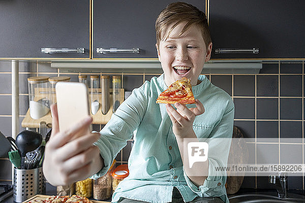 Porträt eines glücklichen Jungen  der in der Küche hausgemachte Pizza isst und sich mit seinem Smartphone selbstständig macht