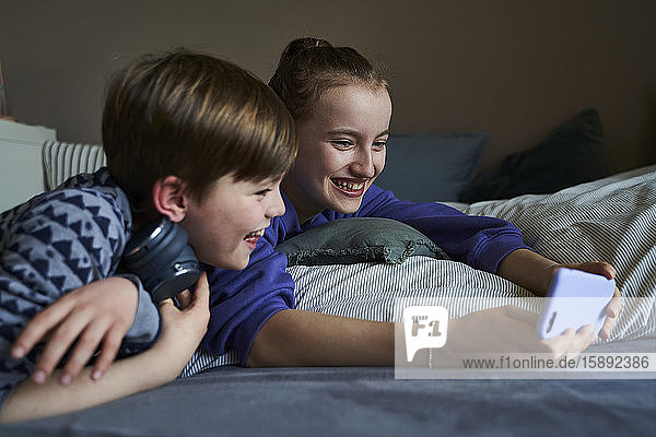 Laughing sibblings lying together on bed using smartphone for video chat