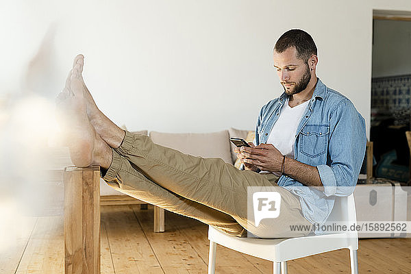 Junger Mann tippt zu Hause mit seinem Smartphone im modernen Wohnzimmer mit den Füßen auf dem Tisch