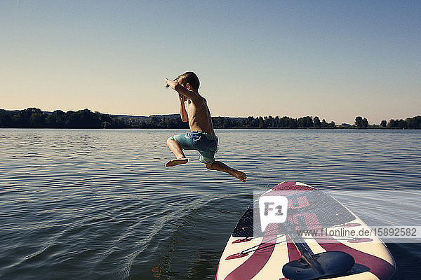 Boy jumping from SUP board into lake at evening twilight