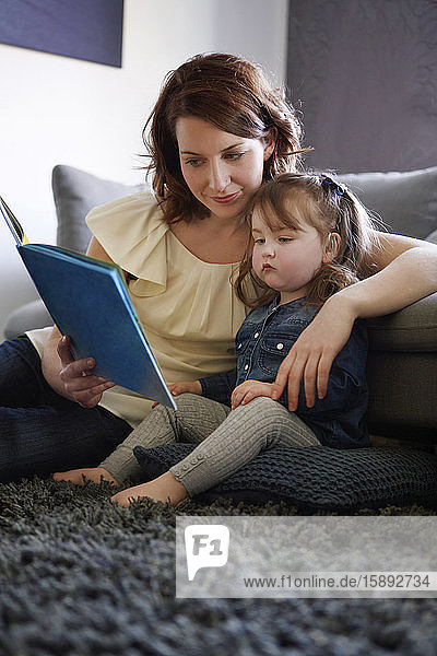Young mother and her little daughter sitting on the floor at home watching picture book