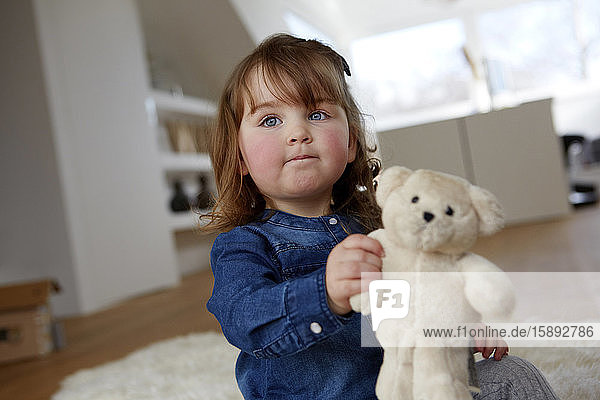 Porträt eines Kleinkindes mit Teddybär  das zu Hause auf dem Boden sitzt