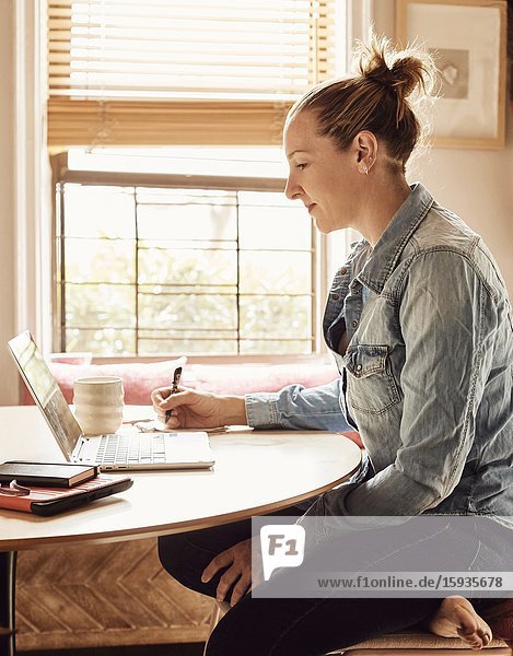 A woman working from home in her sunny kitchen  looking at her computer.