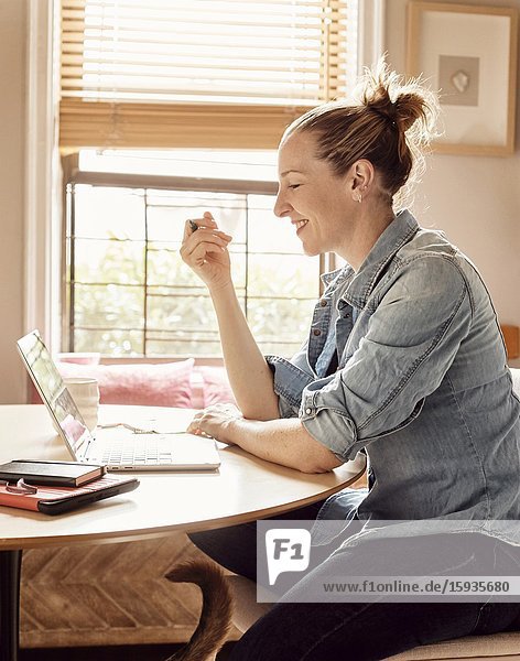 A woman working from home in her sunny kitchen  smiling at the computer.