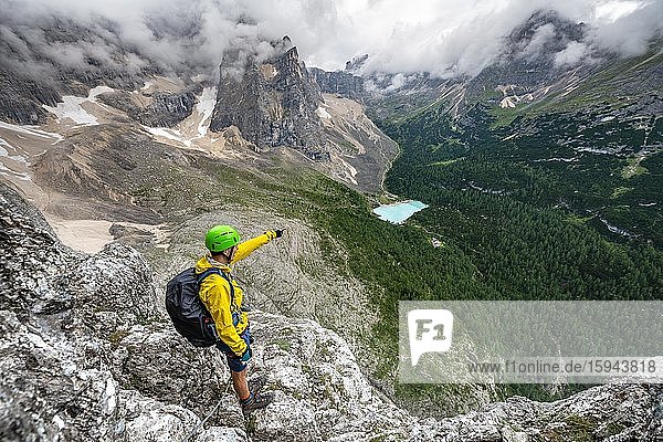 Young man points into the distance  mountaineer on a via ferrata Vandelli  view of Lago di Sorapis  Sorapiss circuit  mountains with low clouds  Dolomites  Belluno  Italy  Europe