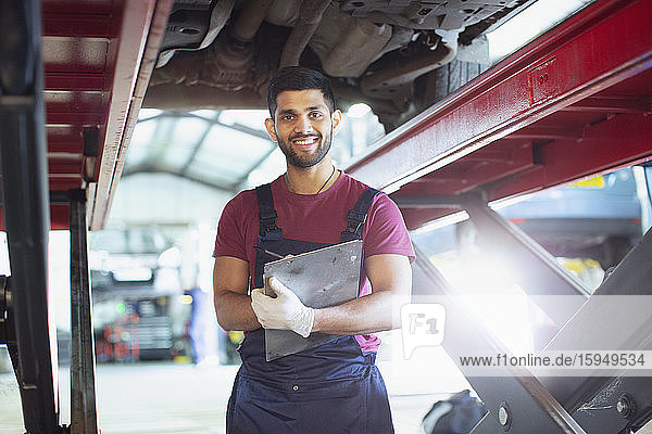 Portrait confident male mechanic with clipboard working under car in auto repair shop