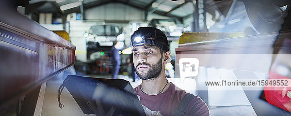 Focused male mechanic with headlight and diagnostic equipment working under car in auto repair shop