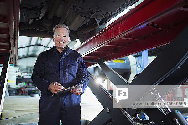 Porträt eines selbstbewussten  lächelnden männlichen Mechanikers mit Klemmbrett  der in einer Autowerkstatt unter dem Auto arbeitet