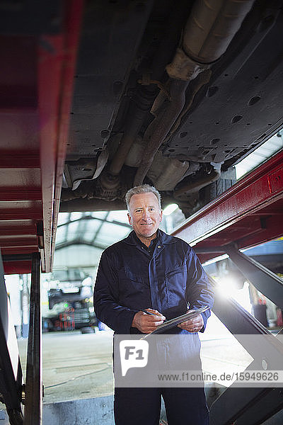 Porträt eines selbstbewussten männlichen Mechanikers mit Klemmbrett  der in einer Autowerkstatt unter dem Auto arbeitet