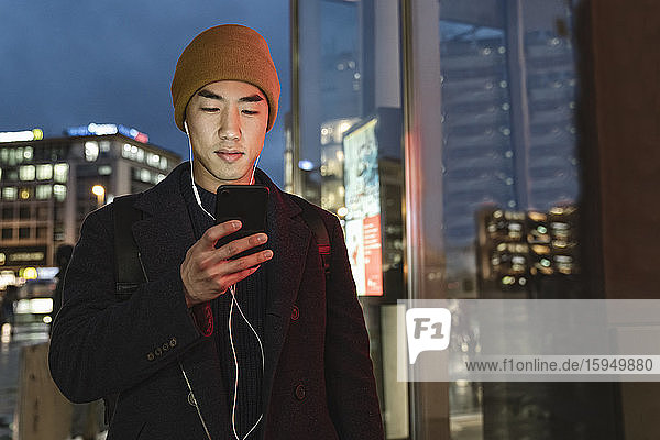 Stylish man with yellow hat and earphones using smartphone at evening on the street at night