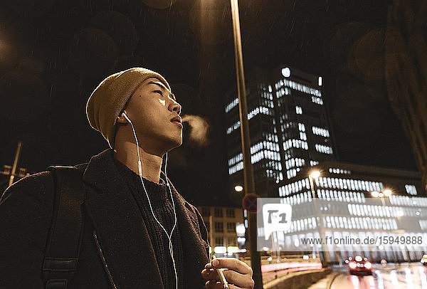 Stilvoller Mann mit gelbem Hut und Kopfhörern  der nachts in der Stadt eine Zigarette raucht